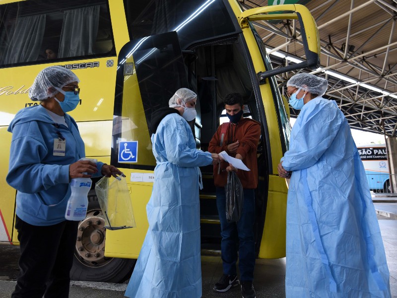 Foto dos agentes de saúde atuando na barreira sanitária do Terminal Rodoviário Tietê. Uma agente com avental azul, touca e máscara afere a temperatura de um rapaz que acabou de descer de um ônibus amarelo. Outras duas agentes auxiliam a ação, uma à direita da foto e outra à esquerda